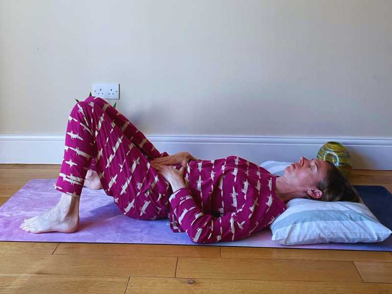 female yoga teacher in pink pjamas lying down on a yoga mat with hands in yoni mudra